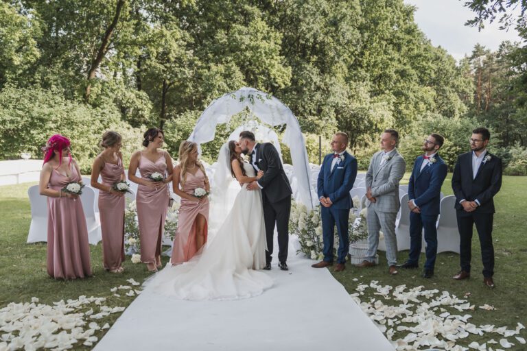 pocałunek pary młodej na tle ceremonii w towarzystwie druhen i drużbów